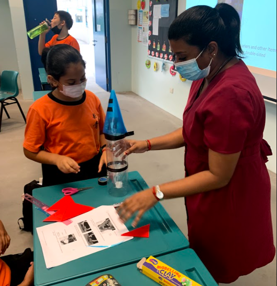 Teacher guides a student wearing a mask in assembling a simple craft or science model using a plastic bottle and paper materials at a classroom table.